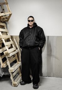 Veston bomber noir avec fermeture éclair, porté sur un sweat à capuche noir. Pantalon noir ample et chaussures foncées. Palettes en bois en arrière-plan.