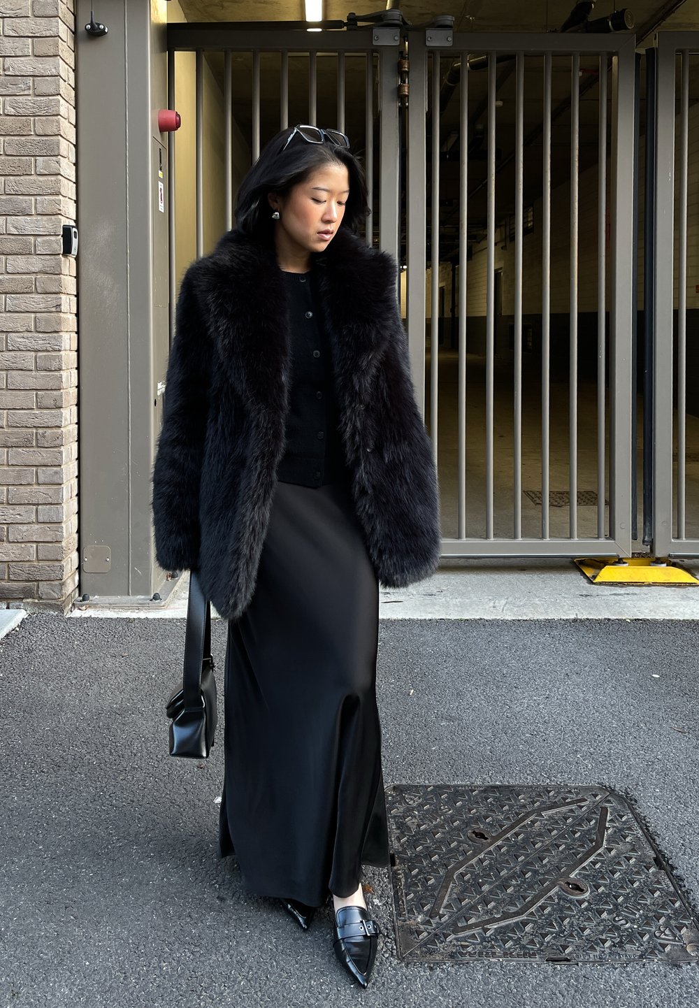 Femme en manteau de fourrure noire et longue jupe marchant sur l'asphalte près d'une porte en métal, tenant un sac à main noir avec des lunettes de soleil sur la tête.