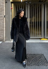 Femme en manteau de fourrure noire et longue jupe marchant sur l'asphalte près d'une porte en métal, tenant un sac à main noir avec des lunettes de soleil sur la tête.