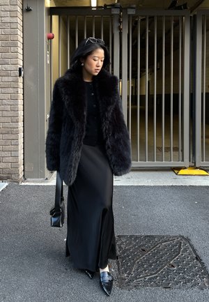 Mujer con abrigo de piel negro y falda larga camina sobre el asfalto cerca de una puerta metálica, sosteniendo un bolso negro y con gafas de sol en la cabeza.