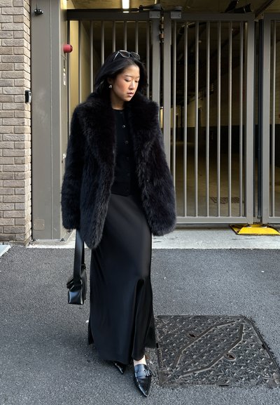Mujer con abrigo de piel negro y falda larga camina sobre el asfalto cerca de una puerta metálica, sosteniendo un bolso negro y con gafas de sol en la cabeza.