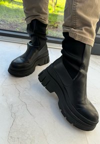 Μαύρες στρατιωτικές μπότες με χοντρή σόλα, λεία δερμάτινη επιφάνεια και ελαστικές πλαϊνές πάνελ, τοποθετημένες σε ένα ανοιχτό, υφασμάτινο πάτωμα.