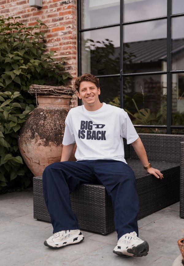 Camiseta blanca con la frase "BIG IS BACK" en letras negras en negrita, combinada con pantalones azul marino y zapatillas blancas, sentada al lado de una vasija de barro.