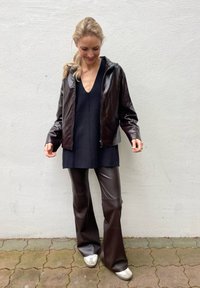 Ciemnobrązowa skórzana kurtka z zamkiem i kapturem, połączona z ciemną bluzką oraz brązowymi spodniami w kształcie dzwonów. Na nogach białe buty z okrągłymi akcentami.