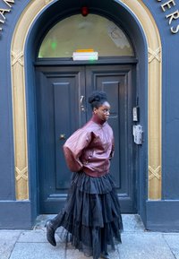 Brązowa skórzana kurtka bomber z dużymi puchowymi rękawami, zestawiona z warstwową czarną spódnicą z tiulu i czarnymi botkami, na tle ciemnoniebieskich drzwi.