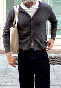 Maglione grigio con bottoni, indossato sopra una camicia bianca. Jeans neri, borsa in cotone e cappellino a fantasia. Ambientazione urbana con pavimento piastrellato.