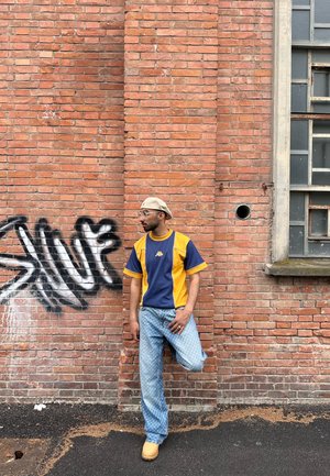 Conjunto informal que incluye una camisa de manga corta azul y amarilla, jeans de un patrón azul claro, y zuecos beige, de pie contra una pared de ladrillo.