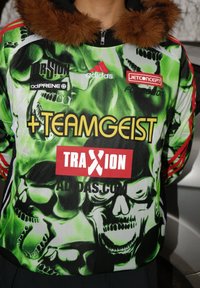 Veste à capuche présentant un imprimé crâne vert vif et noir, avec des accents rouges et un col en fausse fourrure. Plusieurs logos de marque présents.