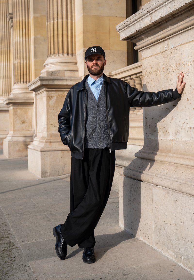 Homme en veste en cuir noire, cardigan gris et casquette noire appuyé contre une colonne en pierre à l'extérieur d'un bâtiment avec de hauts piliers.
