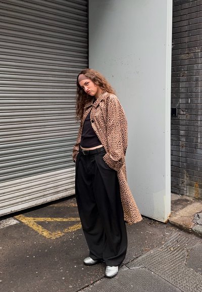 Abrigo largo de estampado de leopardo, top negro, pantalones negros de gran tamaño y zapatos plateados. El fondo presenta una persiana metálica gris y hormigón.
