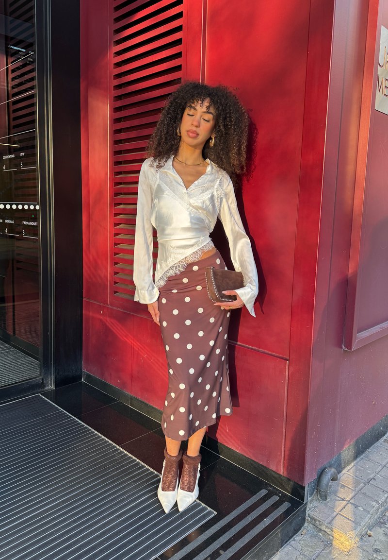 Γυναίκα με σγουρά μαλλιά στέκεται μπροστά σε κόκκινο τοίχο, φοράει λευκό σατέν μπλουζάκι, καφέ φούστα με πουά, καφέ τσάντα και λευκά μυτερά παπούτσια με καφέ κάλτσες.