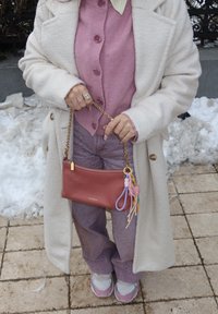 Άτομο που φοράει λευκή ρόμπα, ροζ πουλόβερ με κουμπιά, μοβ παντελόνι με σχέδια και λευκά-ροζ αθλητικά παπούτσια, κρατώντας μια κόκκινη τσάντα με πολύχρωμα μπρελόκ.