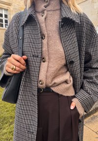Cappotto grigio e bianco a quadri sopra un maglione beige a collo alto, con pantaloni marroni scuri. Borsa nera, cintura in oro opaco e dettagli anello visibili.