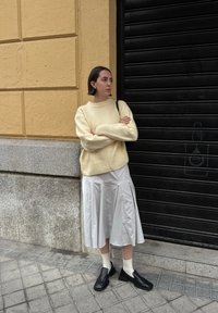 Suéter de malha bege, saia pregueada cinza, sapatos pretos trançados e meias brancas. Aposicionado contra uma parede amarela texturizada e uma persiana preta.