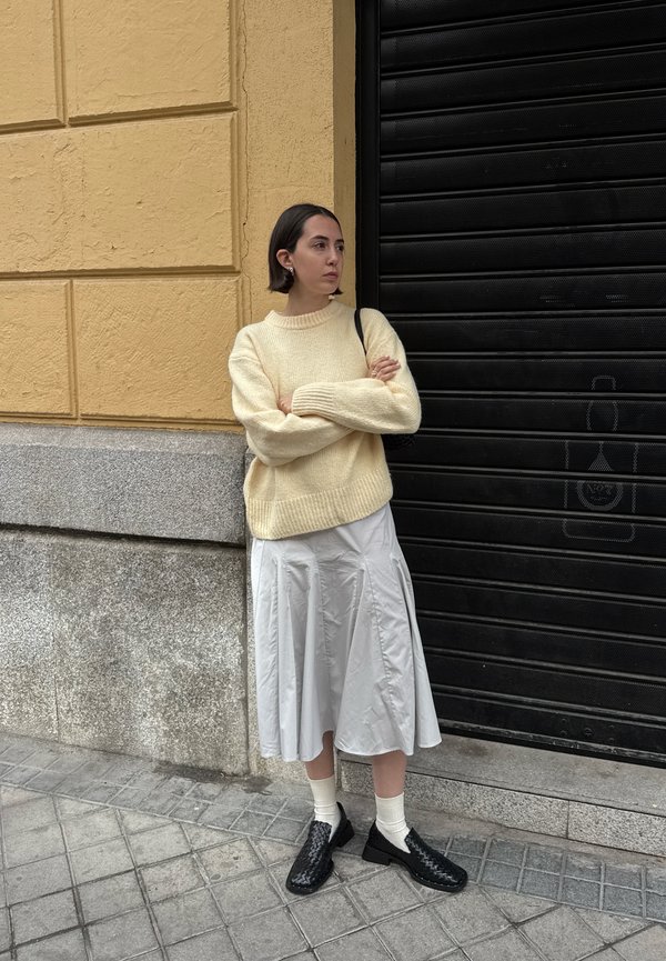 Beige neulepaita, harmaa laskostettu hame, mustat kudotut kengät ja valkoiset sukat. Seisten teksturoidun keltaisen seinän ja mustan ikkunaluukun äärellä.