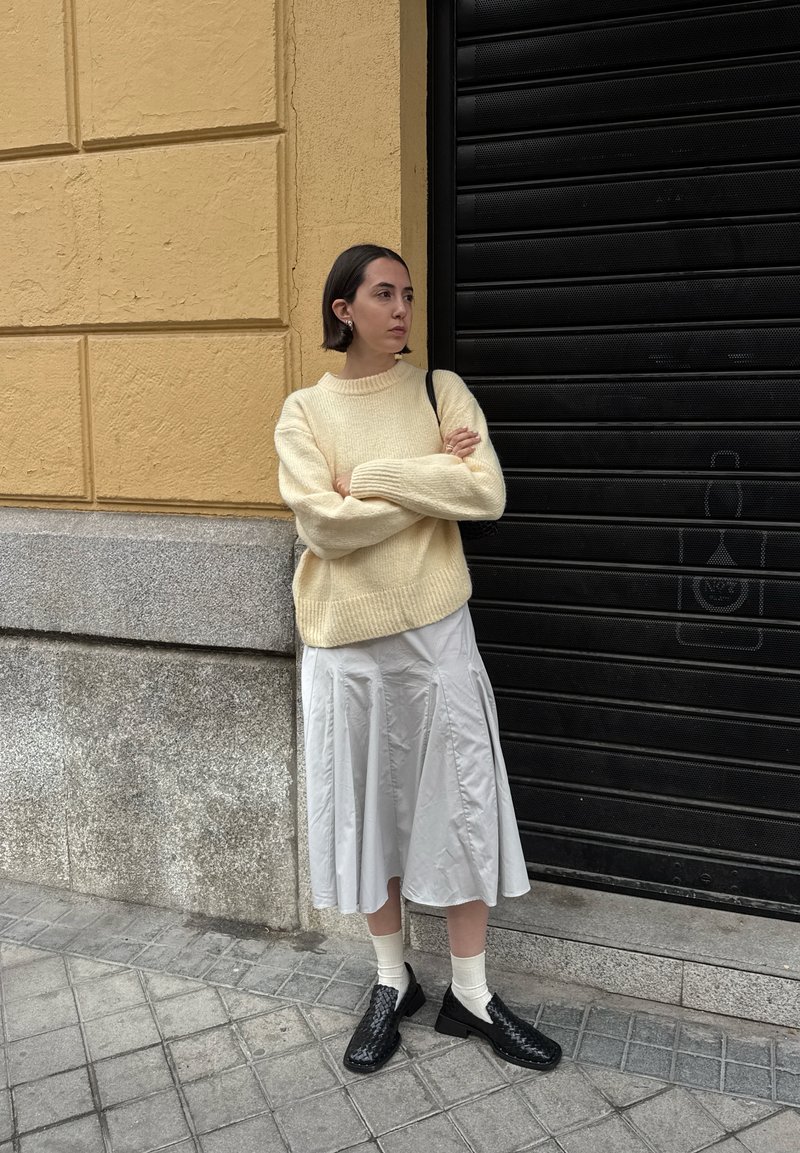 Beiger gestrickter Pullover, grauer Faltenrock, schwarze geflochtene Schuhe und weiße Socken. Steht vor einer strukturierten gelben Wand und einem schwarzen Fensterladen.