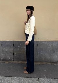 Femme debout de profil portant un béret noir, une veste crème ceinturée, un jean large sombre, des chaussures pointues marron et un sac à bandoulière beige.