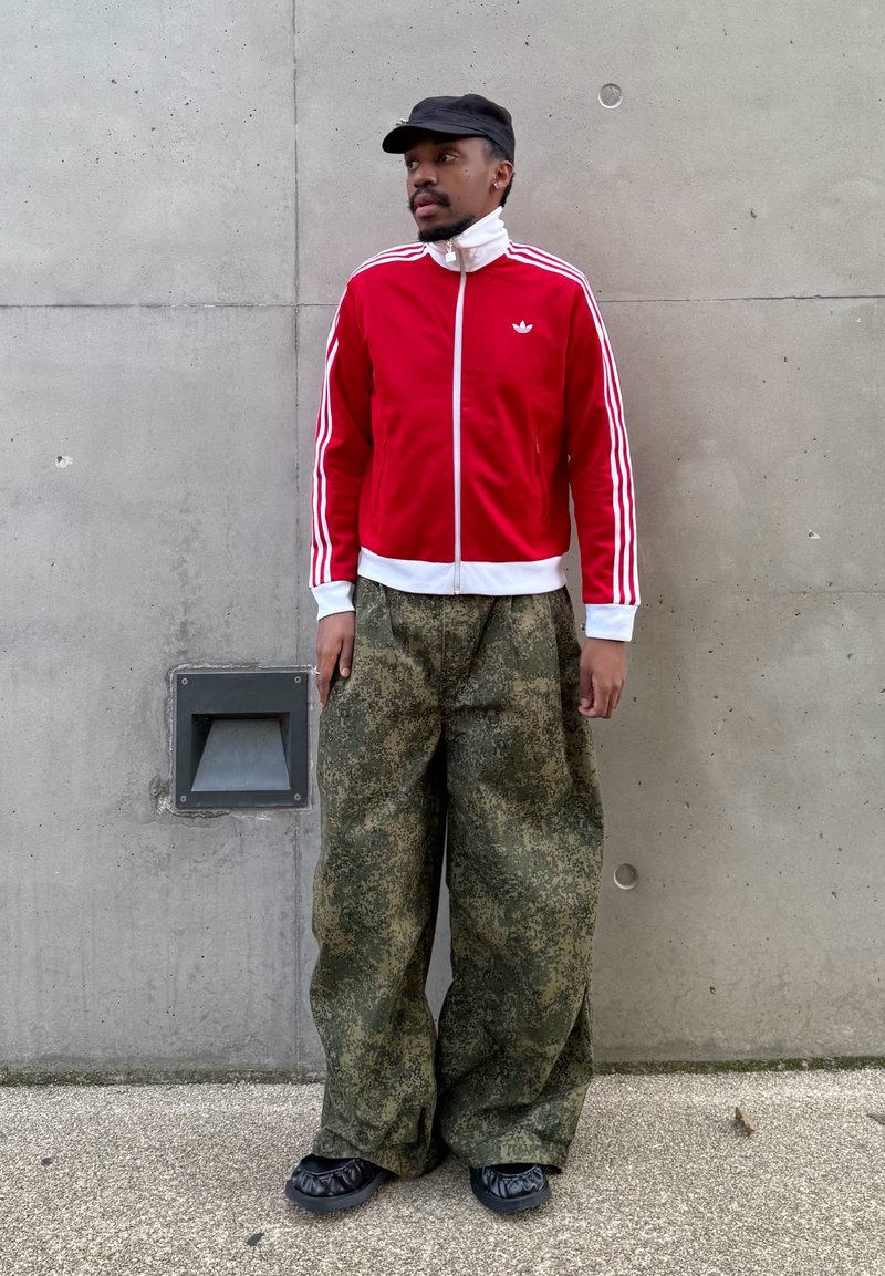 Homme portant une veste de survêtement Adidas rouge, un pantalon large camouflage, une casquette noire et des chaussures noires, debout contre un mur en béton gris.