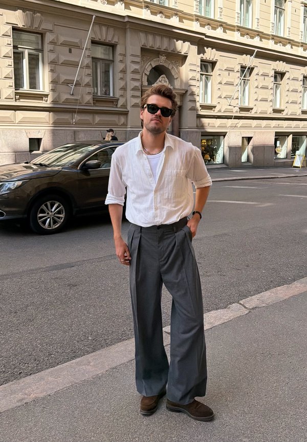 Homme portant des lunettes de soleil, une chemise blanche, un pantalon gris à jambes larges et des chaussures marron, debout dans une rue en ville avec un bâtiment historique et une voiture stationnée derrière.