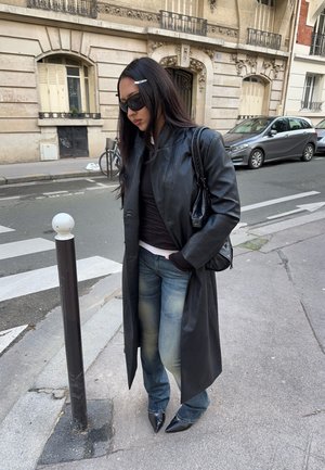 Mujer que lleva un abrigo largo de cuero negro, gafas de sol, vaqueros y zapatos negros puntiagudos, de pie en una calle de la ciudad cerca de un bolardo.