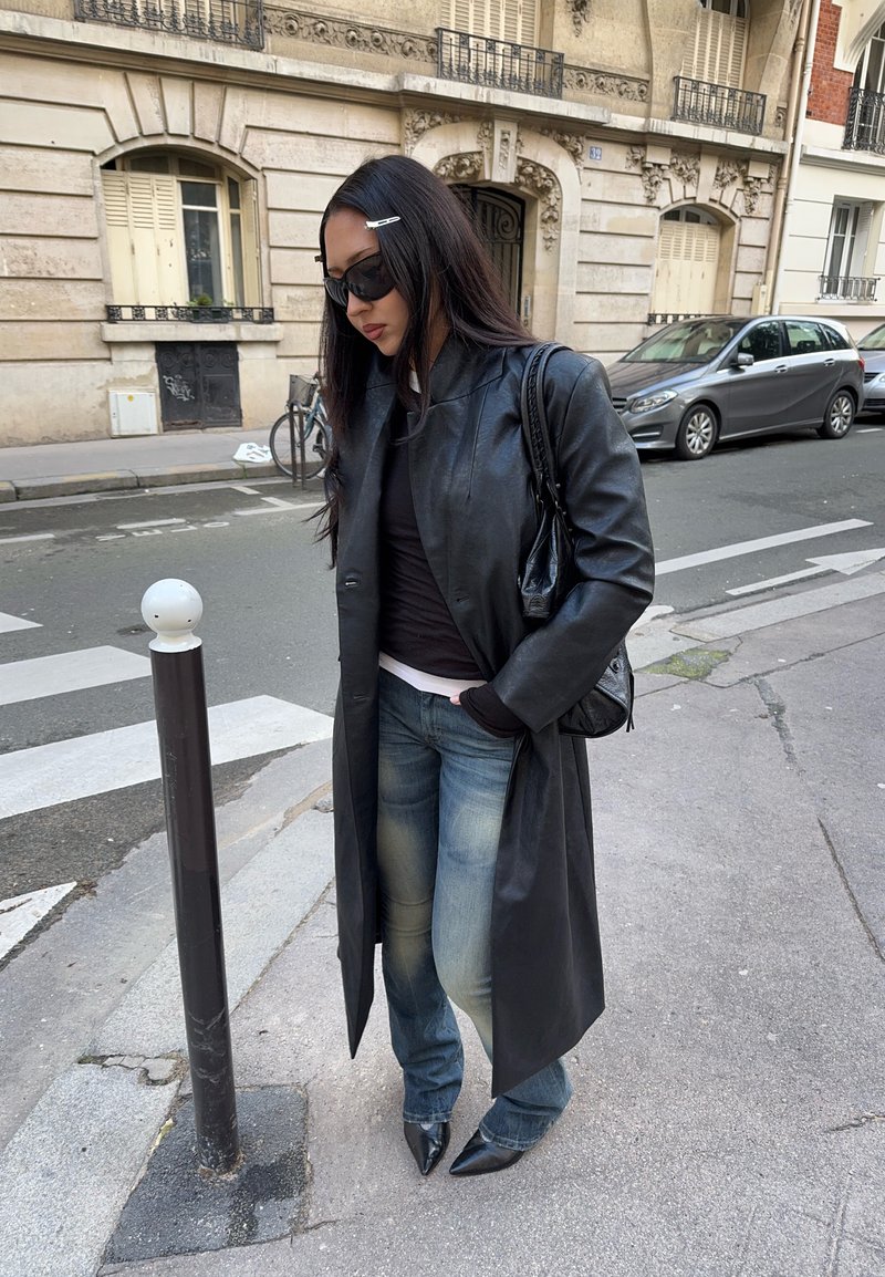 Femeie purtând o haină lungă din piele neagră, ochelari de soare, blugi și pantofi negri cu vârf ascuțit, stând pe o stradă din oraș lângă un stâlp de protecție.