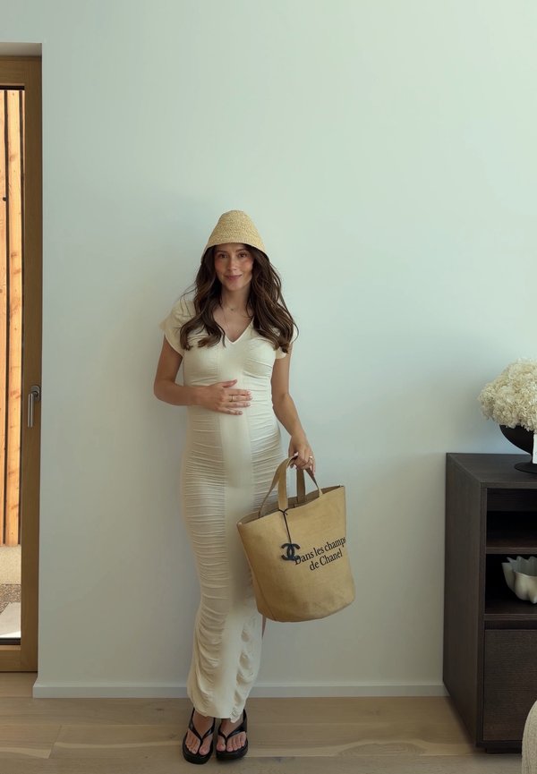 Woman in a beige dress and hat holds a tan Chanel bag, standing indoors by a white wall next to a dark cabinet with a flower arrangement.