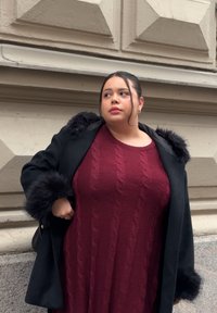 Femme portant une robe en maille torsadée bordeau et un manteau noir avec garniture en fourrure, debout contre un mur en pierre beige, regardant sur le côté.