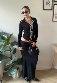Top de manga longa preto com nervuras e detalhes brancos, combinado com uma saia preta esvoaçante. Acessórios incluem uma bolsa preta e um colar de pérolas.