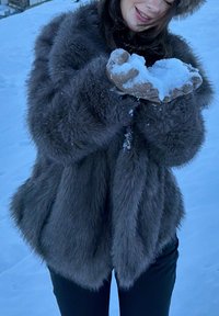 Personne portant un manteau en fourrure gris et un chapeau tenant une poignée de neige à l'extérieur par une journée enneigée, souriant avec les dents visibles.