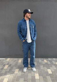 Jeansjacke mit zwei Vordertaschen, getragen über einem hellgrauen T-Shirt, kombiniert mit locker sitzenden Jeans und grauen Turnschuhen, vor einer grauen Wand.