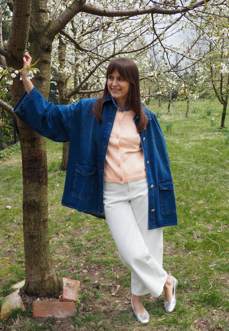 Femme en veste bleue et pantalon blanc appuyée contre un arbre en fleurs dans un verger vert, souriante et regardant sur le côté.