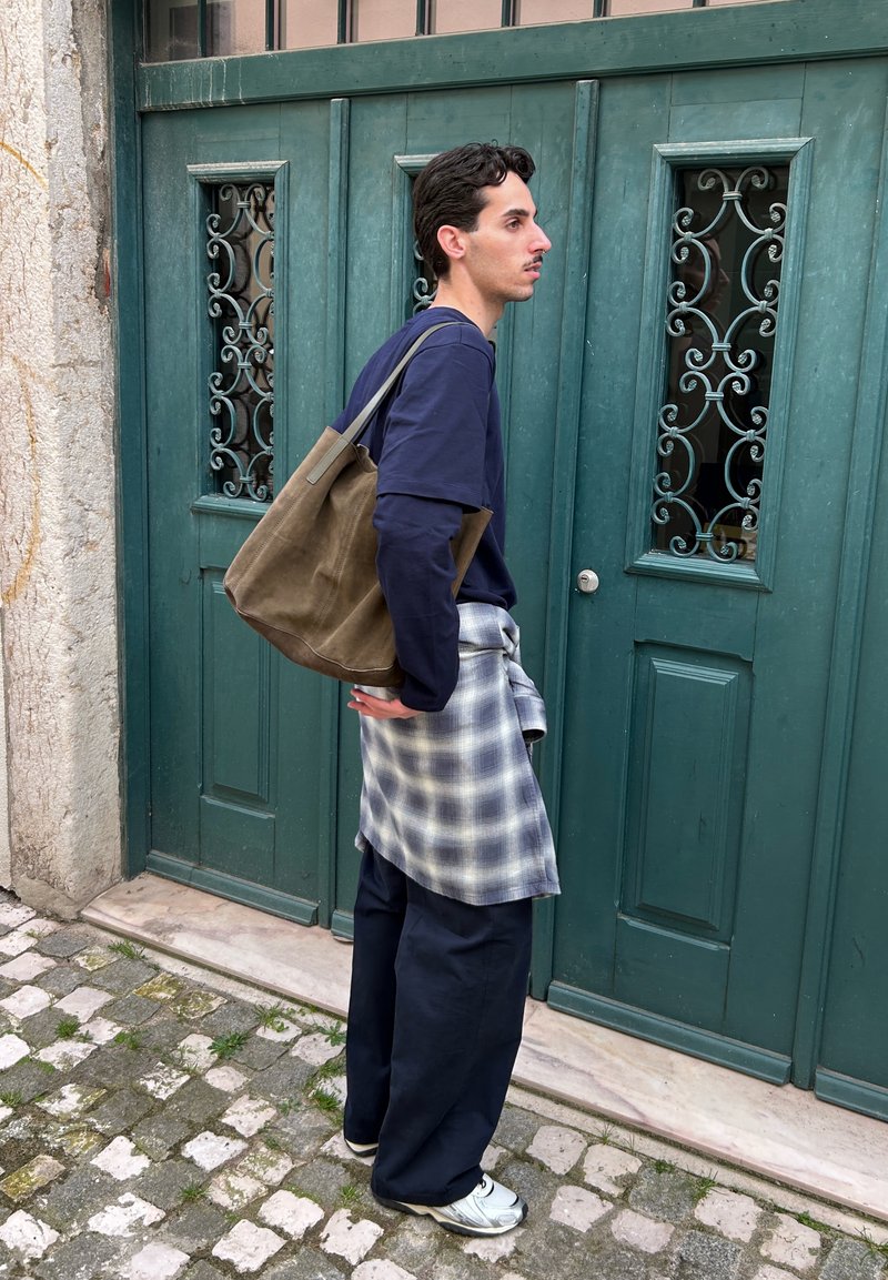 Νέος άνδρας με ναυτικό σύνολο και καρό πουκάμισο δεμένο στη μέση, κρατώντας καφέ τσάντα tote, στέκεται σε λιθόστρωτο δρόμο δίπλα σε πράσινη πόρτα.