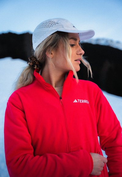 Chaqueta de forro polar rojo con cremallera frontal completa y logo "TERREX", combinada con una gorra blanca perforada. Pelo recogido en una coleta con una goma.