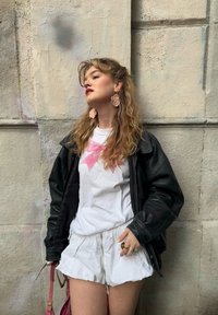 Giacca di pelle nera, t-shirt bianca con fiocco rosa, pantaloni corti bianchi e orecchini floreali. Lo sfondo è una parete in pietra chiara e testurizzata.