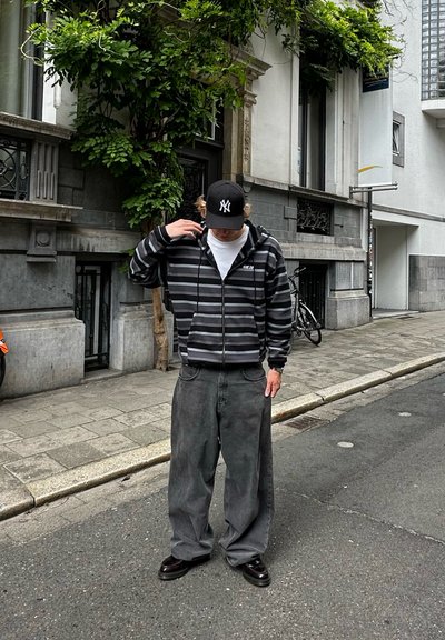 Sudadera a rayas negras y grises con cremallera frontal, jeans grises holgados y zapatos negros; lleva una gorra negra con un logo blanco. Entorno urbano.