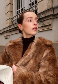 Faux fur jas in bruintinten, met een zachte textuur en brede revers. Gecombineerd met een donkere coltrui en een witte handtas. Gouden oorbellen zichtbaar.