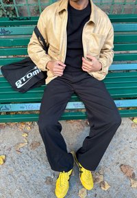 Veste en similicuir beige avec poignets élastiques, pull en maille noire, pantalon large noir, et baskets jaunes à rayures noires. Sac cabas noir posé près des pieds.