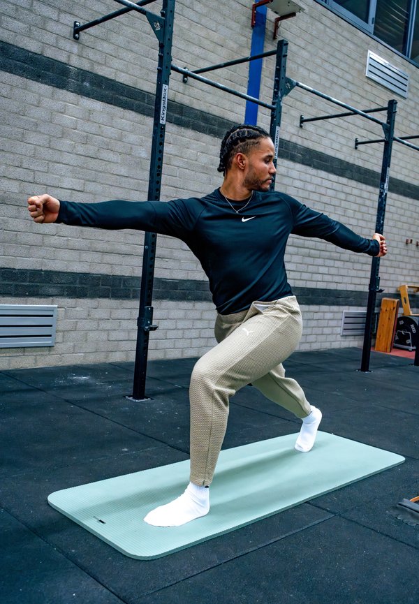 Una persona demuestra una postura de yoga sobre una esterilla de color verde claro, vistiendo una camiseta de manga larga negra y pantalones texturizados en tono arena, en un entorno de gimnasio.
