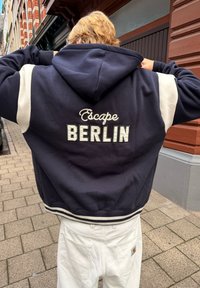Persona rubia con una sudadera azul marino con el texto "Escape BERLIN" en la parte posterior y pantalones blancos, de pie en una acera de la ciudad.