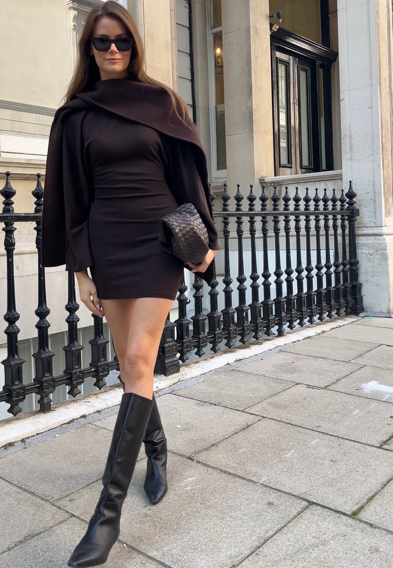 Form-fitting black dress with long sleeves, paired with a dark wrap and knee-high black boots. Accessories include a woven black clutch.