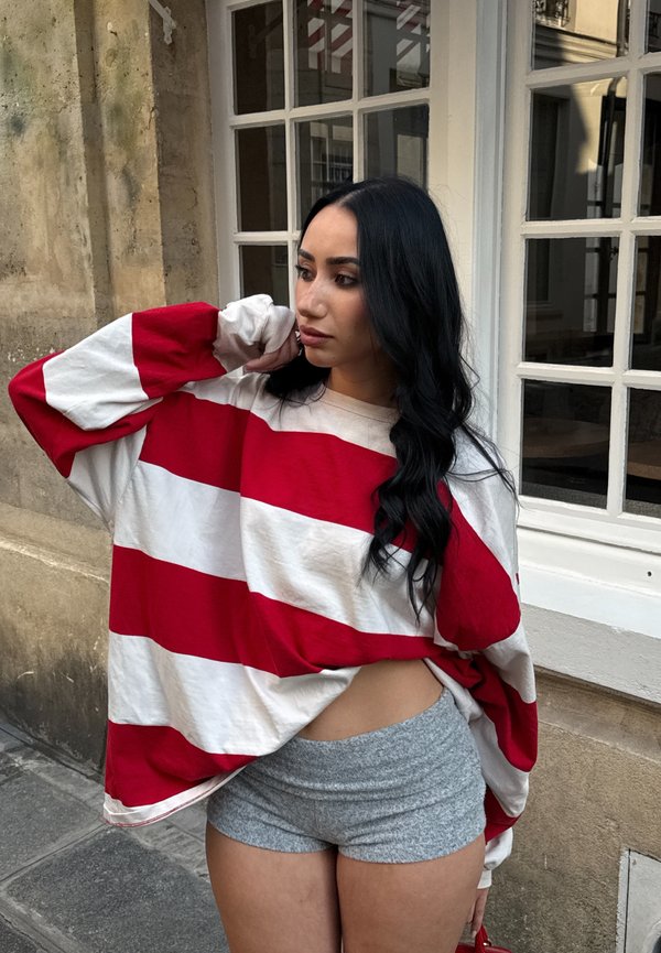 Mujer con cabello largo y negro que lleva una camisa holgada a rayas rojas y blancas y pantalones cortos grises, de pie cerca de un edificio con ventanas, mirando hacia un lado.
