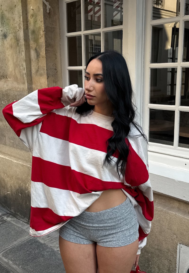 Femme aux longs cheveux noirs portant une chemise ample rayée rouge et blanche et un short gris, debout près d'un bâtiment vitré, regardant sur le côté.