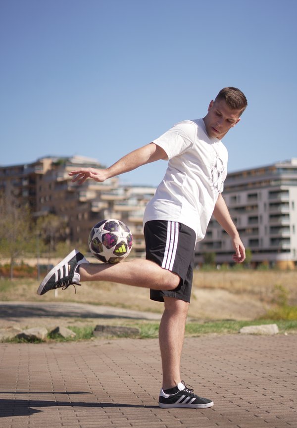 Joven con camisa blanca y pantalones cortos negros equilibrando un balón de fútbol en su pie levantado al aire libre con edificios al fondo.