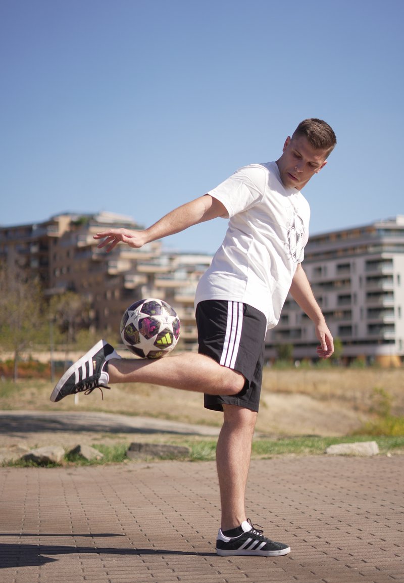 Jeune homme en chemise blanche et short noir équilibrant un ballon de football sur son pied levé en plein air avec des bâtiments en arrière-plan.