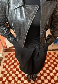 Zwarte bikerjack van imitatieleren met een getextureerde afwerking, een cropped ontwerp en ritssluiting, gecombineerd met losse zwarte broek en puntige schoenen.