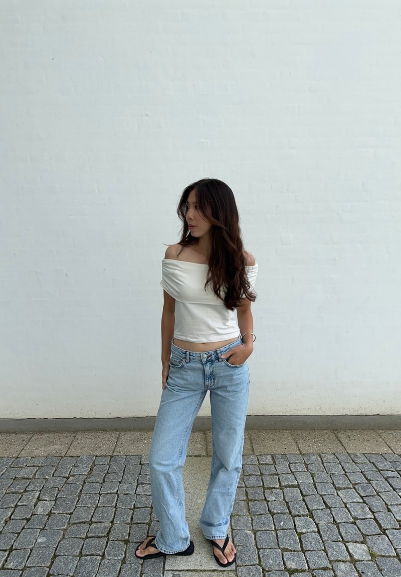 Κρέμα λαιμόκοψη μπλούζα και ανοιχτό μπλε τζιν παντελόνι. Η μπλούζα έχει μαζεμένη υφή; το τζιν είναι φαρδύ και με ίσια γραμμή. Μαύρες παντόφλες.