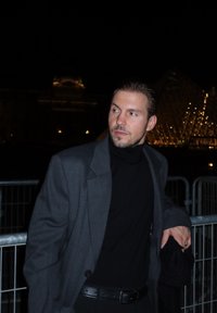 Grijze blazer over een zwarte coltrui, donker haar naar achteren gekamd, staand bij metalen barrières met een verlichte architecturale achtergrond.