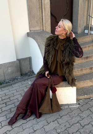 Brown satin trousers, olive green blouse with a bow, faux fur gilet, and a brown bag featuring a rose accent. Long black lace sleeves. Square-toed boots.