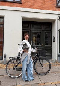 Melnas krāsas velosipēds ar grozu, viegls pelēks džemperis saistīts ap baltu krekliņu un platām gaiši zilām džinsa biksēm. Fons no ķieģeļu un baltas sienas.