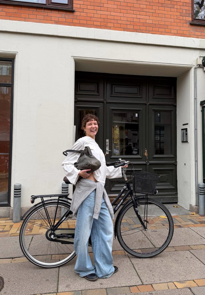 Bicicleta preta com um cesto, suéter cinza leve atado sobre uma blusa branca, e jeans largos azul claro. Fundo de parede em tijolo e branco.